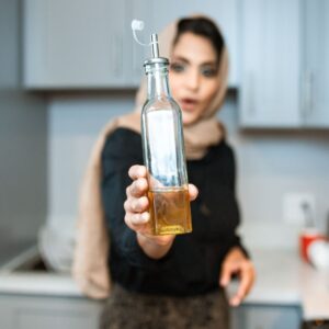 Positive ethnic woman wearing headscarf showing glass bottles of organic olive or sunflower oil while cooking in modern kitchen