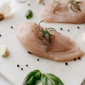 Close-up of uncooked chicken breasts with rosemary, basil, and spices on a white board.
