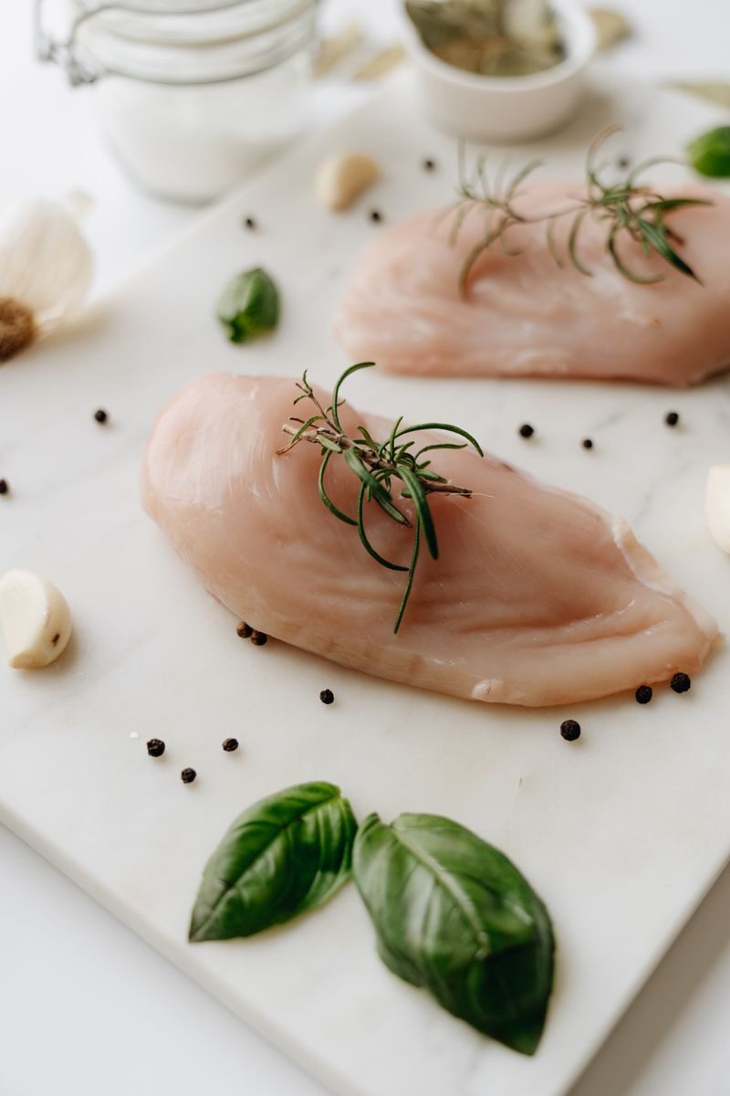 Close-up of uncooked chicken breasts with rosemary, basil, and spices on a white board.